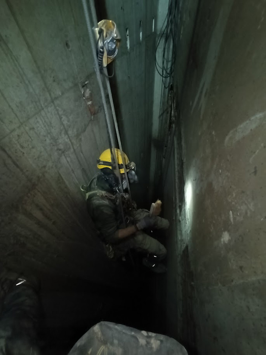 Technicien cordiste équipé d'un masque respiratoire complet en rappel dans un conduit industriel étroit et sombre.