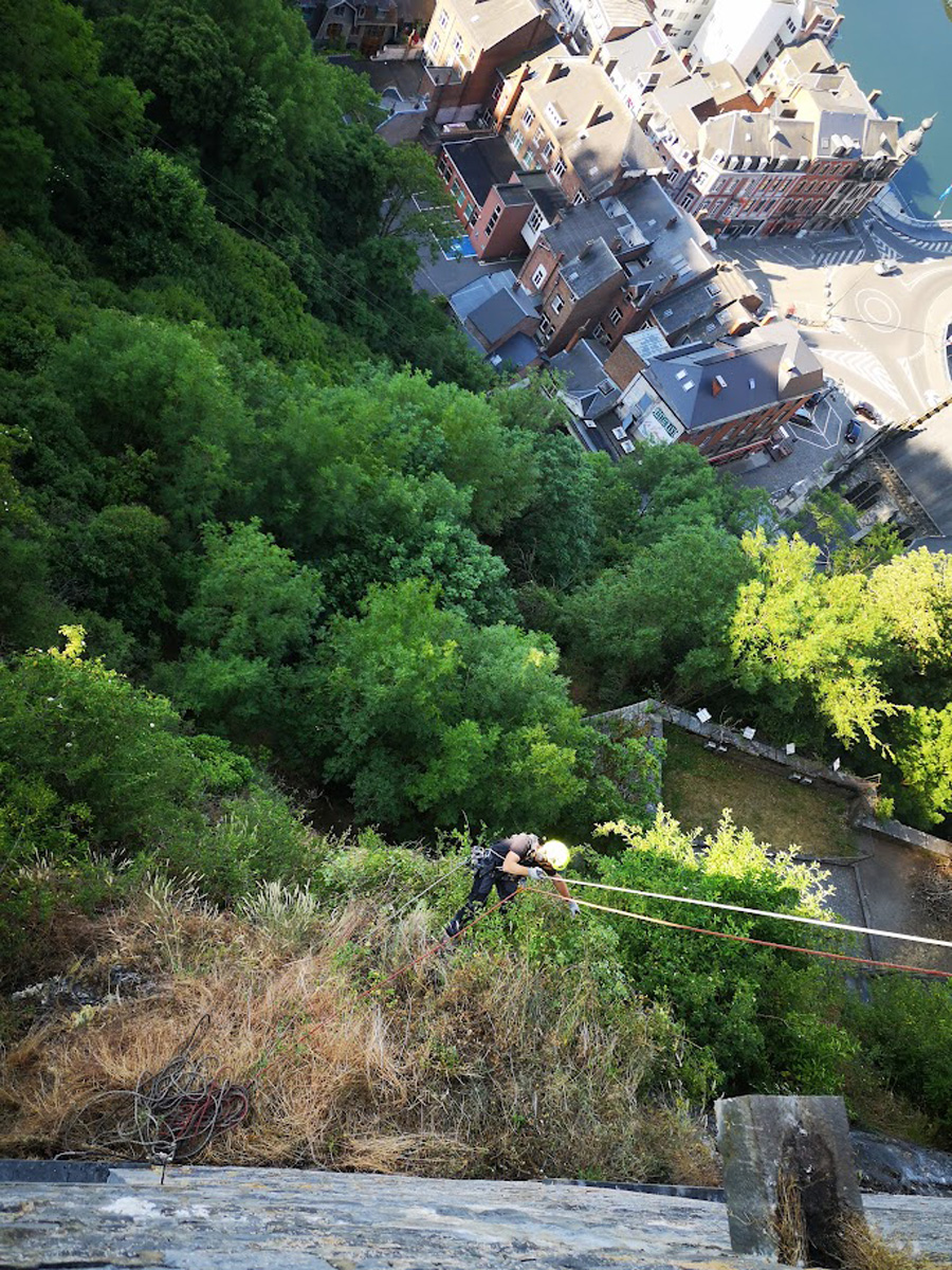 Intervention technique de sécurisation et de purge de falaise par un cordiste expert Vertikal Pro sur une paroi rocheuse surplombant une vallée fluviale.