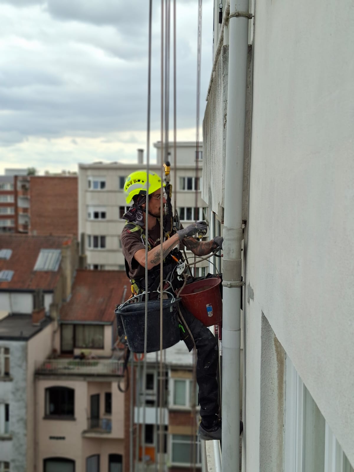 Un cordiste en tenue de sécurité, portant un casque jaune fluorescent et suspendu à des cordes, travaille sur la façade d’un immeuble en hauteur. Il utilise des outils près d’une canalisation, avec des seaux de matériel attachés à son équipement. À l’arrière-plan, on aperçoit des immeubles résidentiels sous un ciel nuageux.
